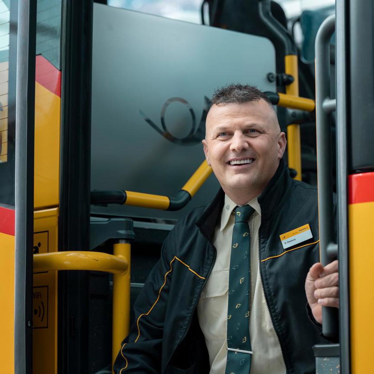 Ein Postautochauffeur vor seinem Postauto, der in die Kamera lacht